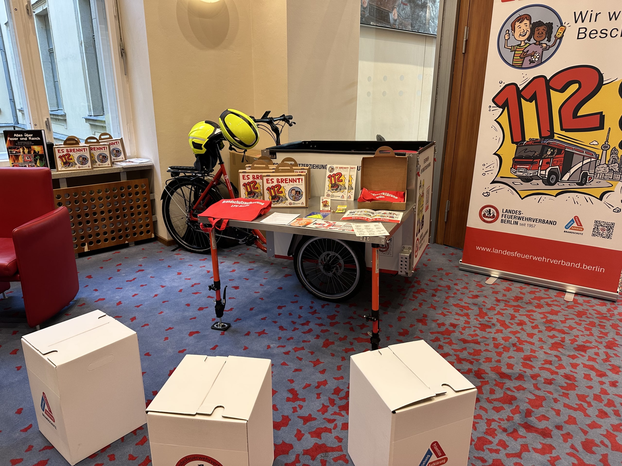 Ein Foto des Feuerwehrinformationsfahrrads im aufgebauten Zustand in einem Raum. Vor dem Fahrrad sind Sitzhocker aufgebaut und das Fahrrad hat den Tisch aufgebaut auf dem Infomaterial und ein Kamishibai Erzähltheater aufgebaut stehen. Es sind keine Personen auf dem Foto.
